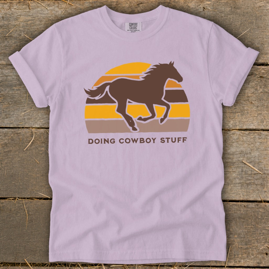 Lilac t-shirt with brown horse silhouette and striped sunset graphic that says "DOING COWBOY STUFF".