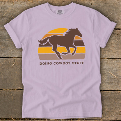 Lilac t-shirt with brown horse silhouette and striped sunset graphic that says "DOING COWBOY STUFF".