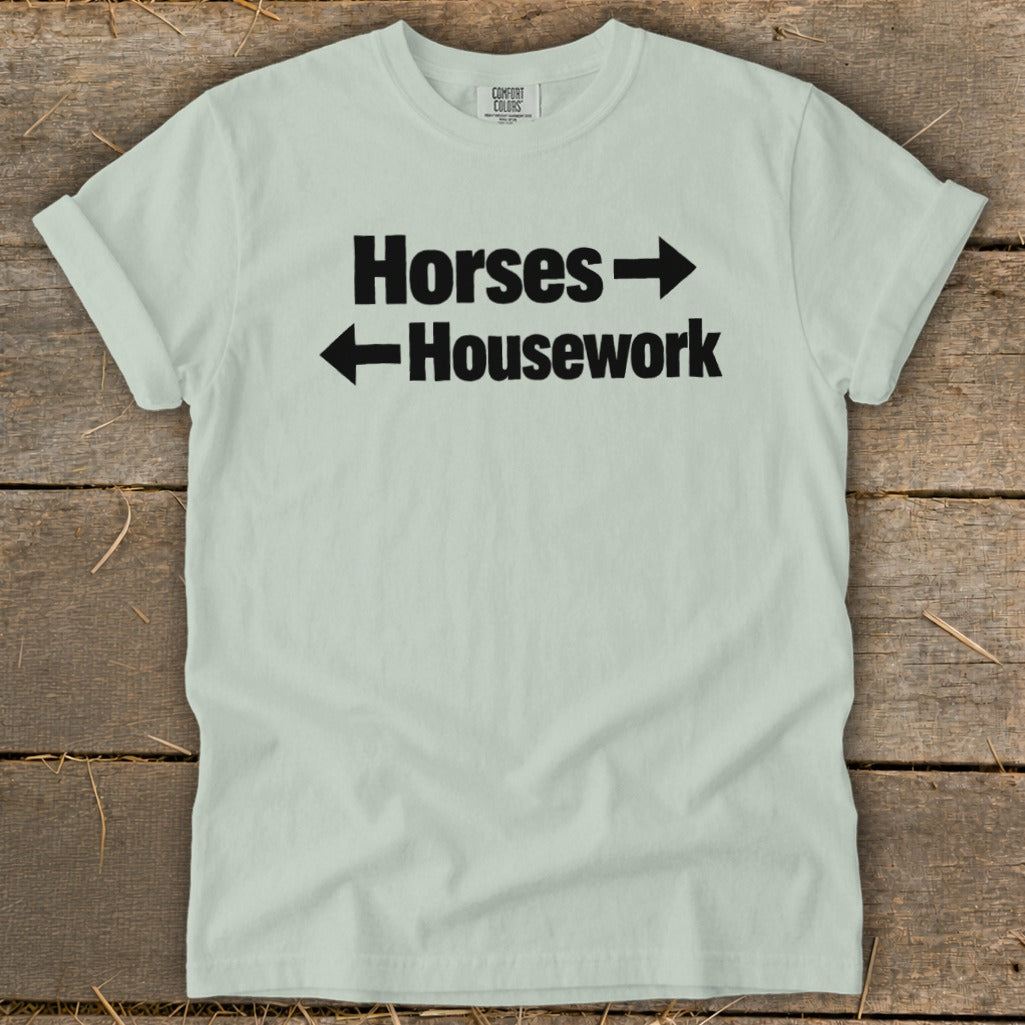 A light green t-shirt with black text that reads Horses with a right arrow above Housework with a left arrow.