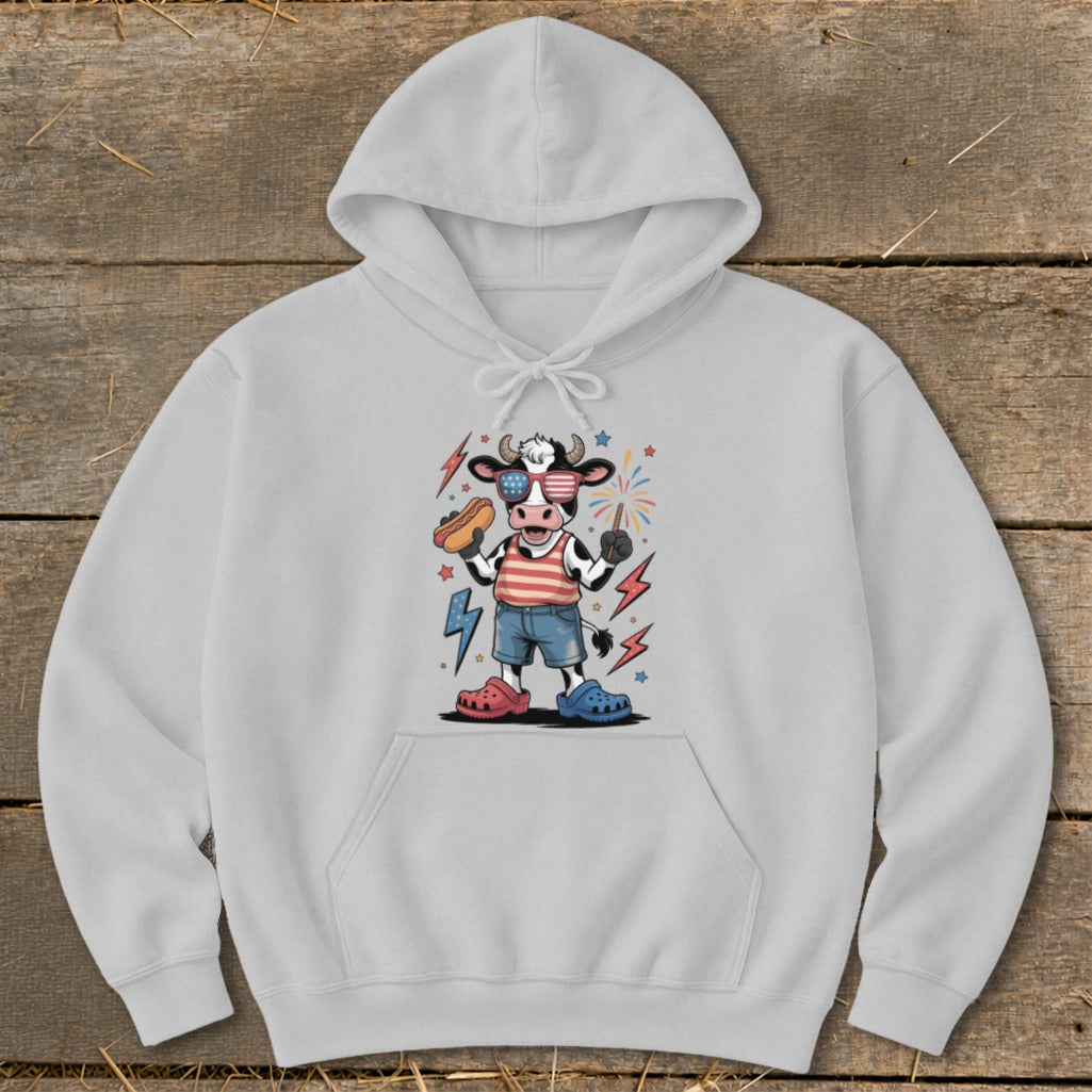Cartoon cow in patriotic sunglasses holding a hot dog and a sparkler.