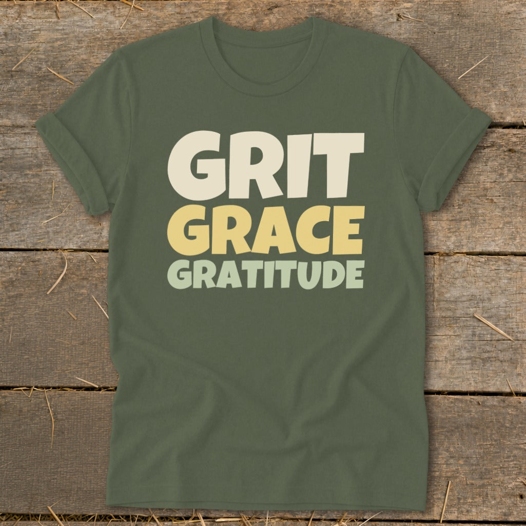 Olive green crewneck t-shirt with rolled sleeves laid flat on a rustic wooden surface, featuring the stacked white and yellow text "GRIT GRACE GRATITUDE".