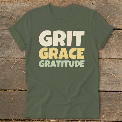 Olive green crewneck t-shirt with rolled sleeves laid flat on a rustic wooden surface, featuring the stacked white and yellow text "GRIT GRACE GRATITUDE".