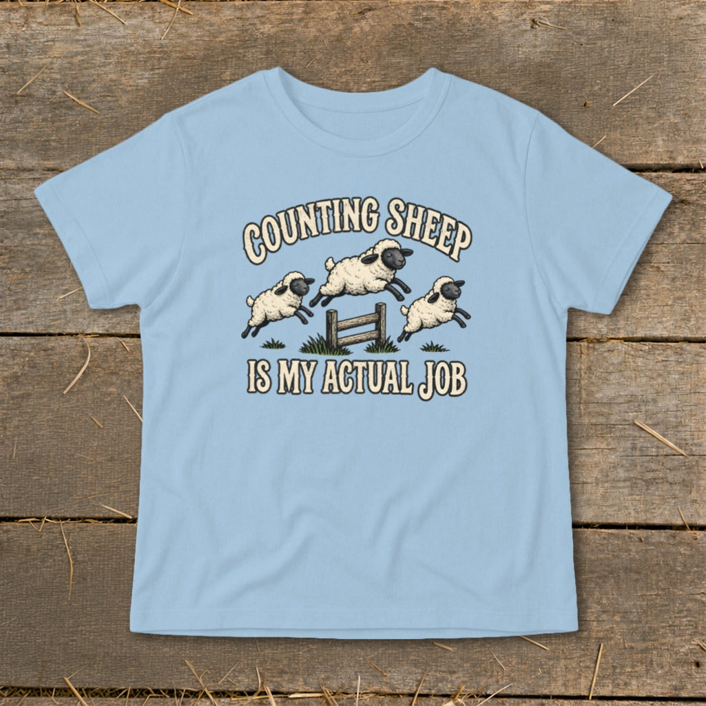 Light blue t-shirt with graphic of three sheep jumping over a fence and text "COUNTING SHEEP IS MY ACTUAL JOB".