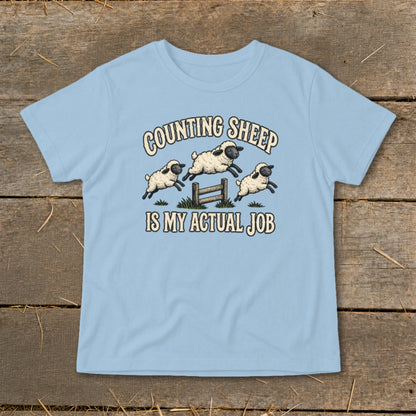 Light blue t-shirt with graphic of three sheep jumping over a fence and text "COUNTING SHEEP IS MY ACTUAL JOB".