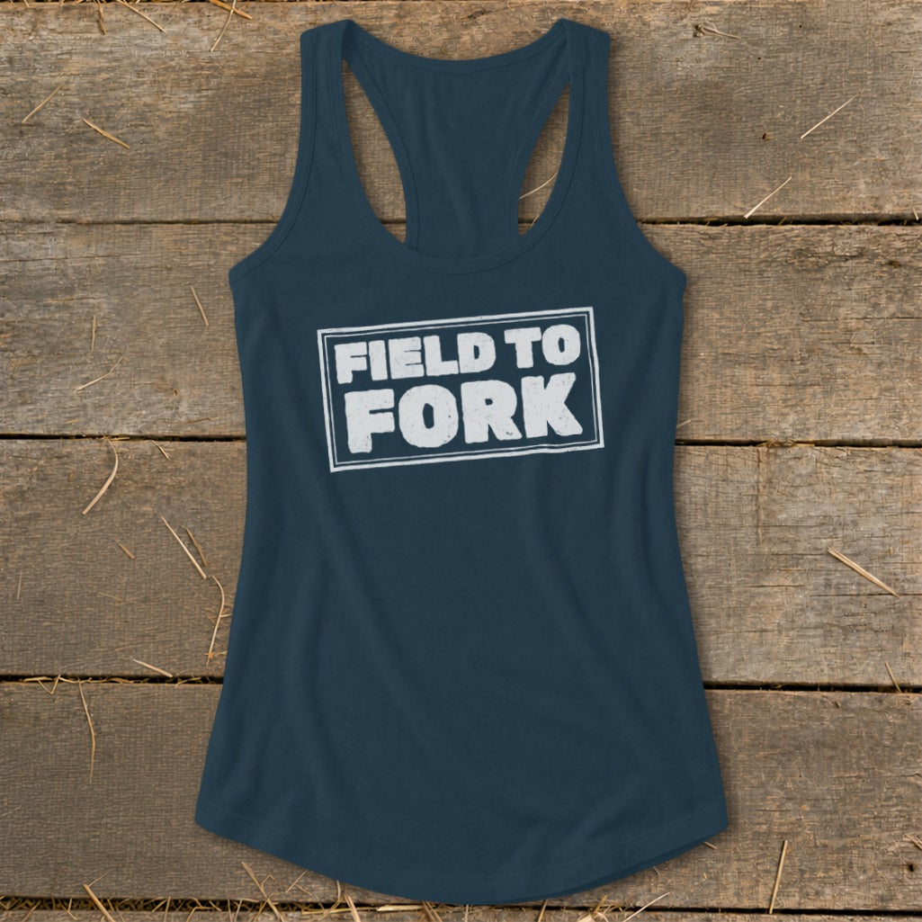 Dark blue racerback tank top featuring the white distressed text FIELD TO FORK laid out flat on a rustic wooden background.