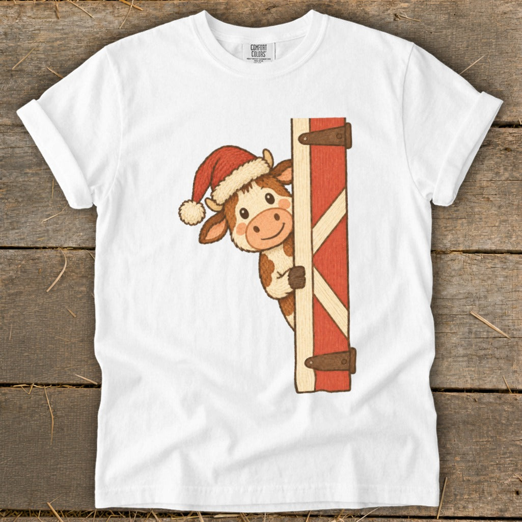 Cartoon cow in Santa hat peeking from behind a barn door on a white t-shirt.