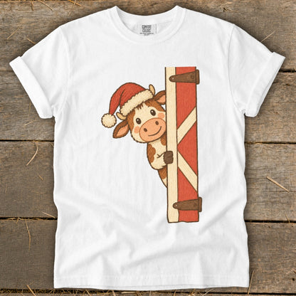 Cartoon cow in Santa hat peeking from behind a barn door on a white t-shirt.