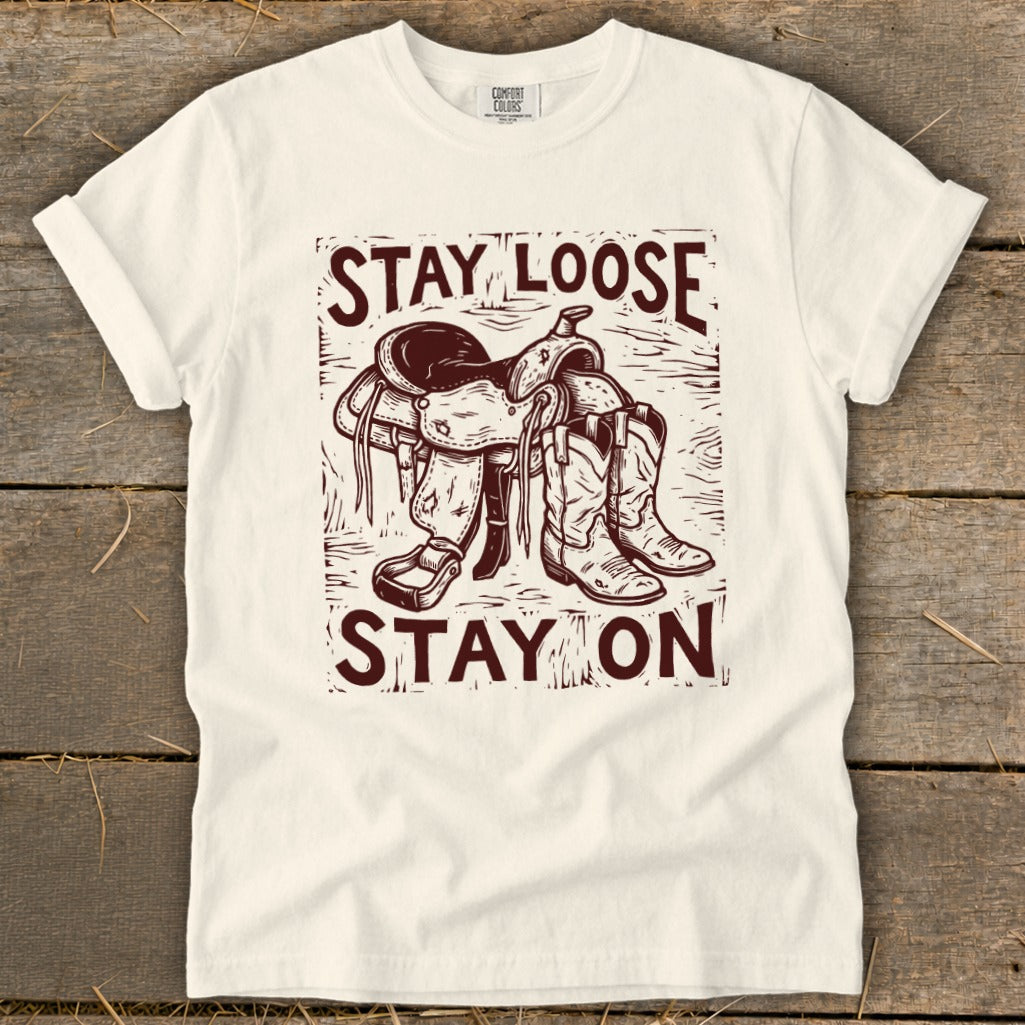 White t-shirt graphic with saddle, boots, and text "STAY LOOSE STAY ON".
