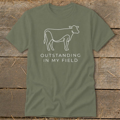 Olive green t-shirt with white line drawing of a cow and text "OUTSTANDING IN MY FIELD".