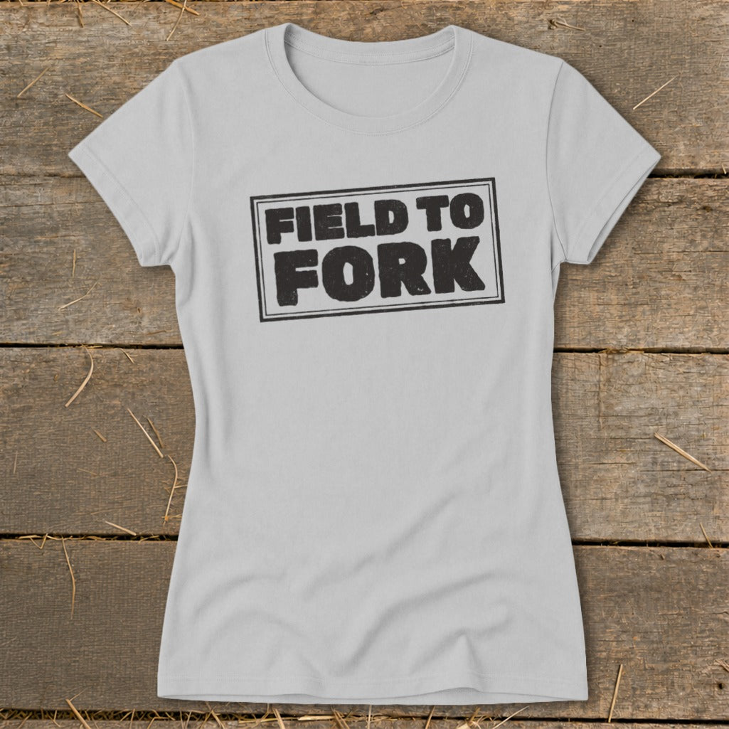Light gray women's t-shirt featuring a black box graphic with the text "FIELD TO FORK" on a rustic wooden background.