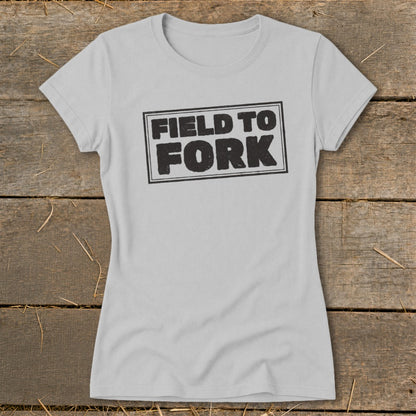 Light gray women's t-shirt featuring a black box graphic with the text "FIELD TO FORK" on a rustic wooden background.
