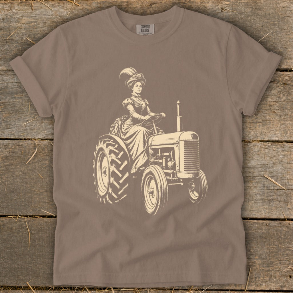 Woman in vintage dress and hat driving a tractor, with "COMFORT COLORS" logo.