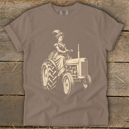 Woman in vintage dress and hat driving a tractor, with "COMFORT COLORS" logo.