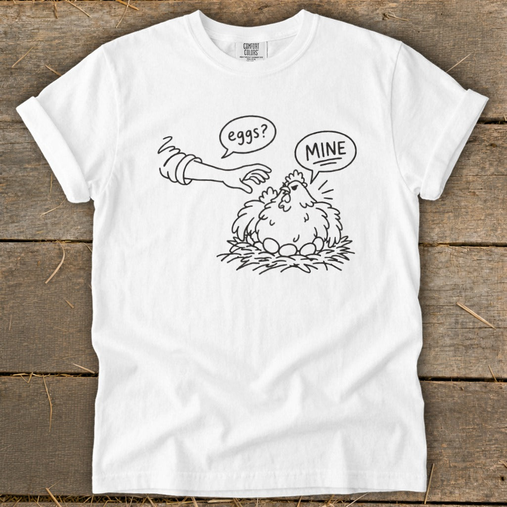 White t-shirt with cartoon chicken on nest saying "MINE" as hand reaches for eggs.