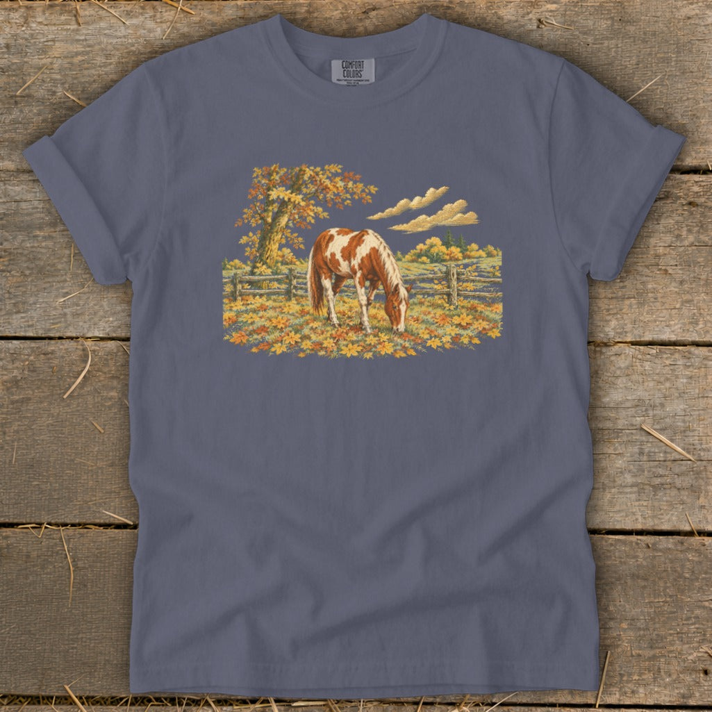 Brown and white horse grazing in a field of autumn leaves with a fence and tree.