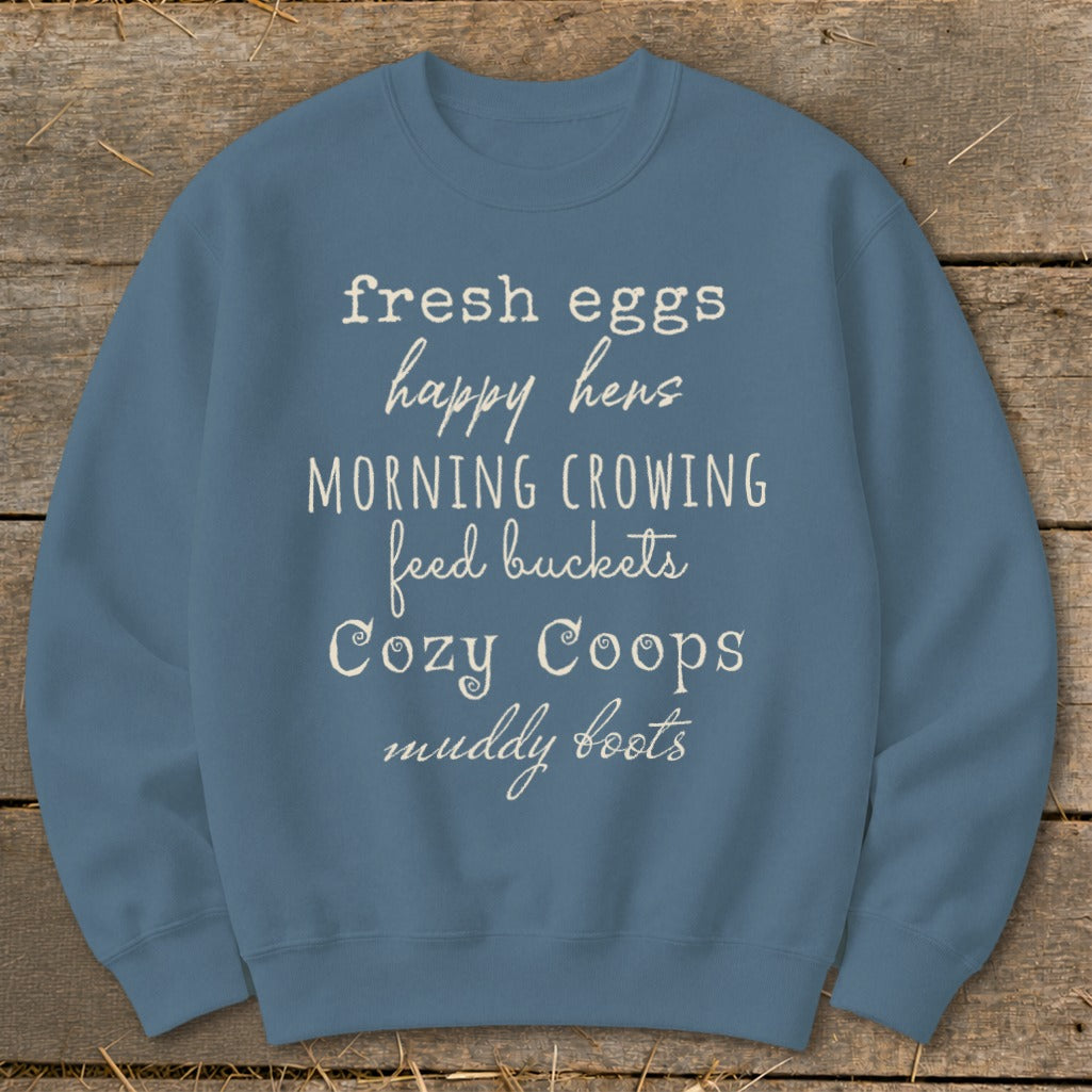 Blue sweatshirt with white text listing "fresh eggs," "happy hens," "morning crowing," "feed buckets," "Cozy Coops," and "muddy boots."