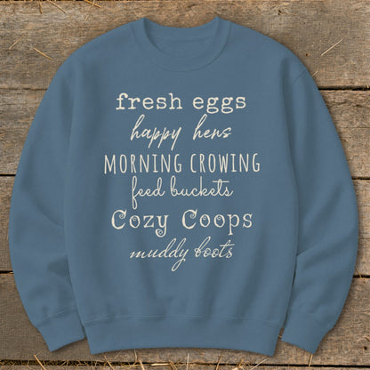 Blue sweatshirt with white text listing "fresh eggs," "happy hens," "morning crowing," "feed buckets," "Cozy Coops," and "muddy boots."