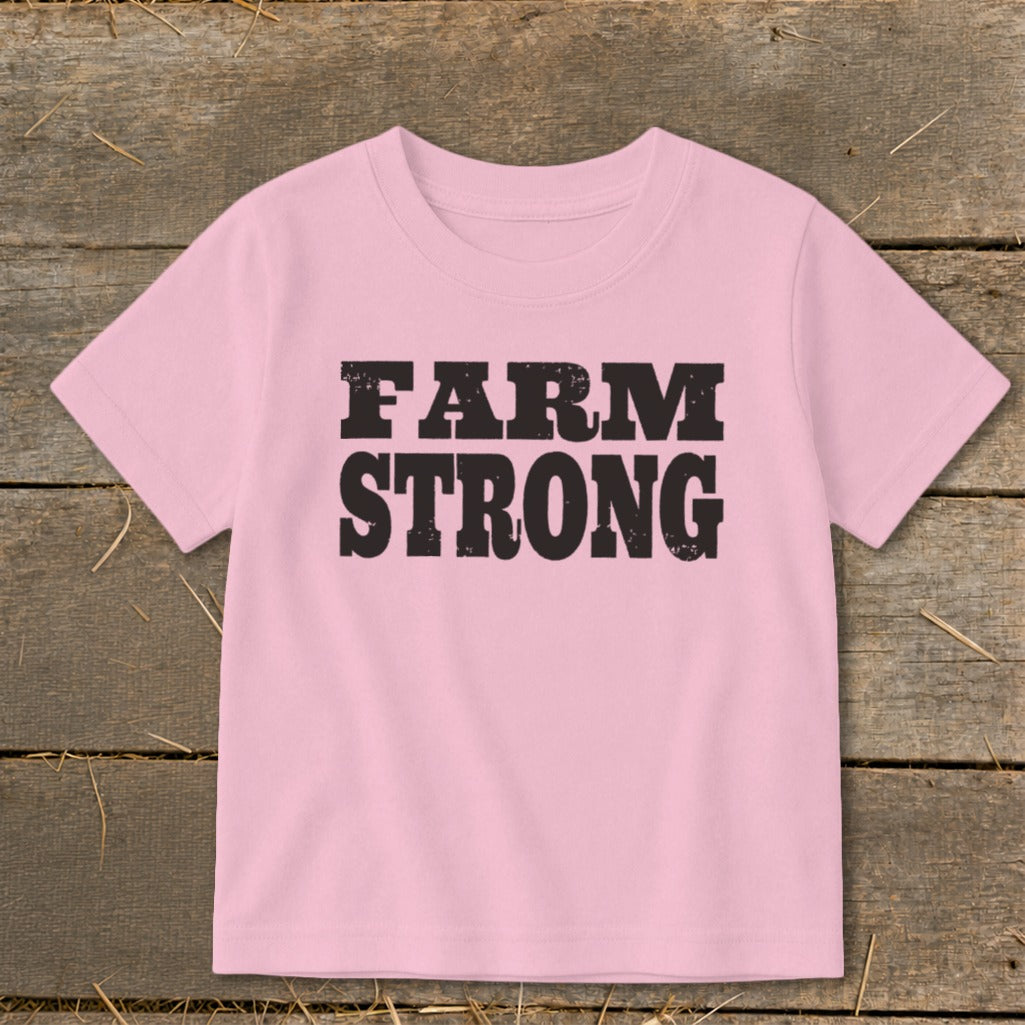 A short-sleeved pink t-shirt with the text FARM STRONG printed in black across the chest, laid flat on a rustic wooden surface.