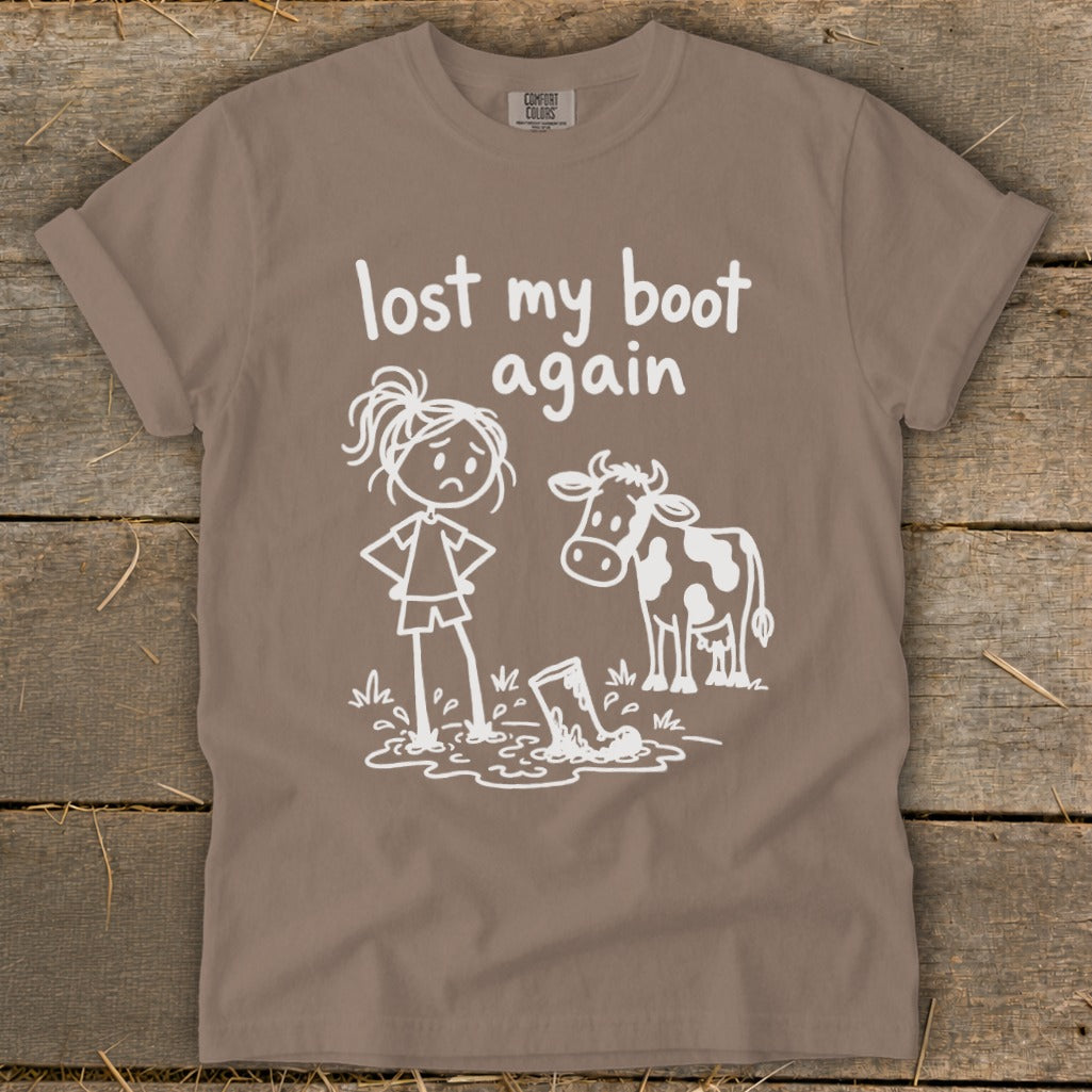 Brown t-shirt featuring white text that reads lost my boot again and a graphic of a sad girl and a cow standing near a boot stuck in mud.