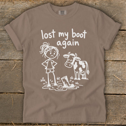 Brown t-shirt featuring white text that reads lost my boot again and a graphic of a sad girl and a cow standing near a boot stuck in mud.