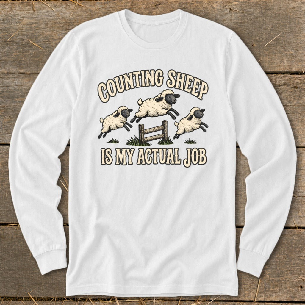 White long-sleeve shirt with sheep jumping over a fence, text "Counting Sheep Is My Actual Job".
