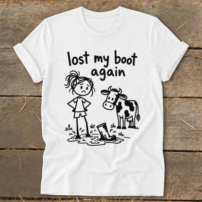 White t-shirt with a black graphic of a sad stick figure standing in mud next to a cow, with the text "lost my boot again".
