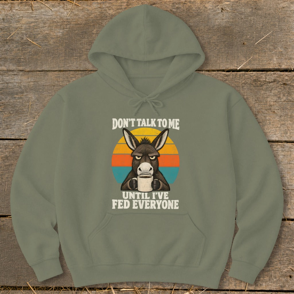 An olive green hoodie featuring a graphic of a grumpy donkey holding a coffee mug with the text Don't Talk to Me Until I've Fed Everyone.