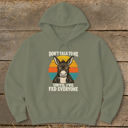 An olive green hoodie featuring a graphic of a grumpy donkey holding a coffee mug with the text Don't Talk to Me Until I've Fed Everyone.