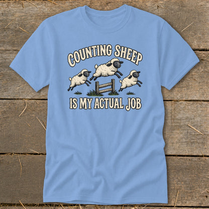 Light blue t-shirt with cartoon sheep jumping over a fence, reading "Counting Sheep Is My Actual Job".