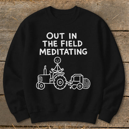Black sweatshirt with white text "OUT IN THE FIELD MEDITATING" and stick figure meditating on tractor.