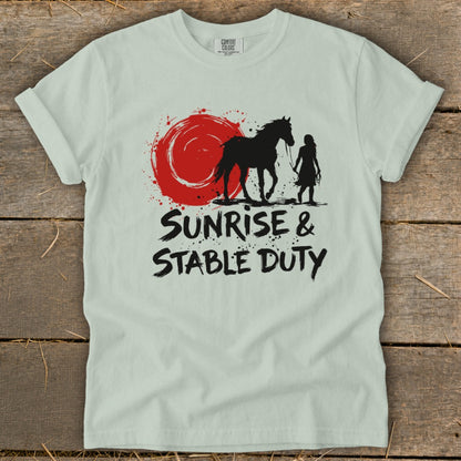 Light green t-shirt with black silhouette of a horse and rider beside a red sun graphic, text reads "Sunrise & Stable Duty."