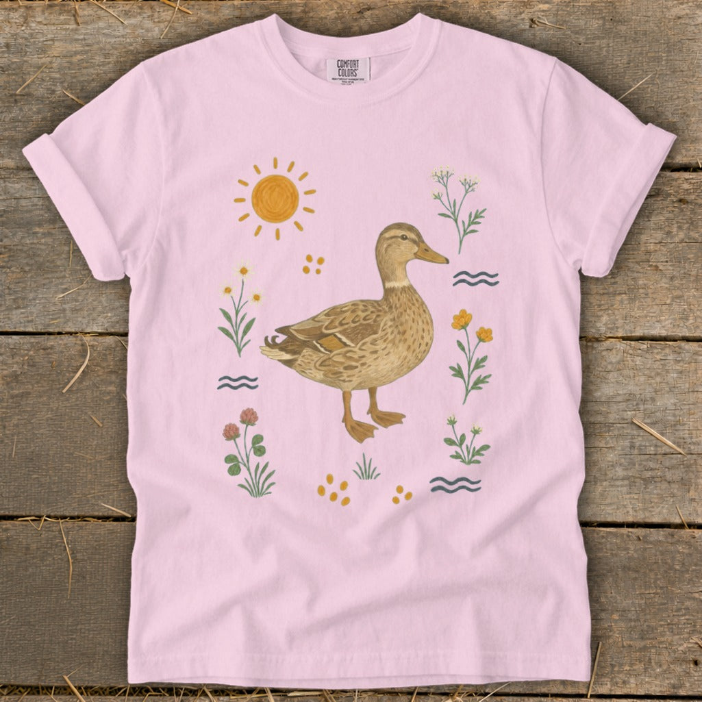 Pink t-shirt with a watercolor duck surrounded by flowers, sun, and water ripples.