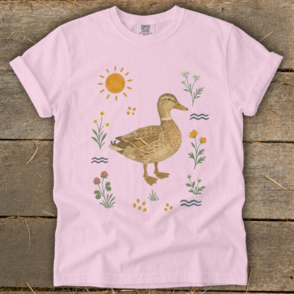Pink t-shirt with a watercolor duck surrounded by flowers, sun, and water ripples.