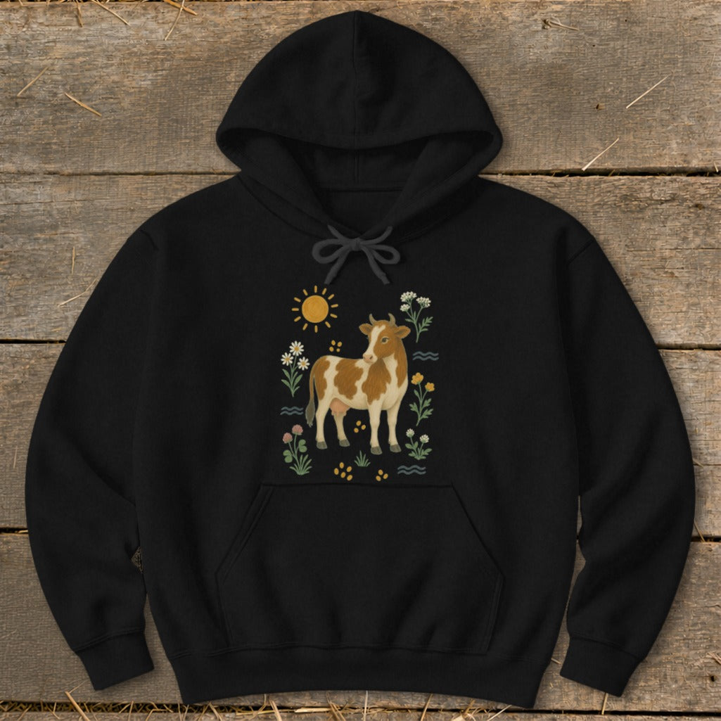 Black hoodie with illustration of a brown and white cow surrounded by sun, flowers, and waves.