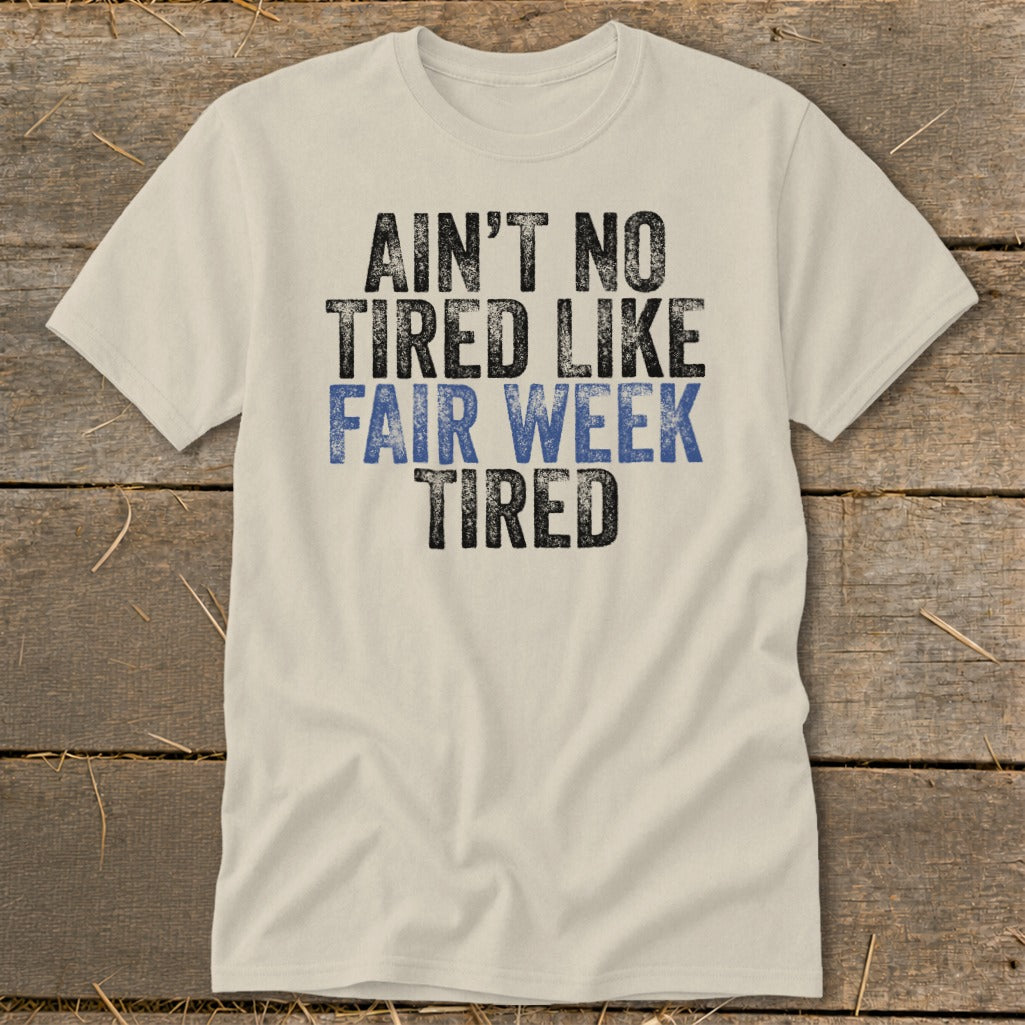 Cream t-shirt with black and blue text reading "AIN'T NO TIRED LIKE FAIR WEEK TIRED".