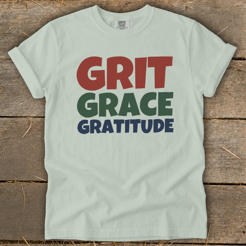 Light green t-shirt laid flat on a rustic wooden background, featuring the words Grit, Grace, and Gratitude.