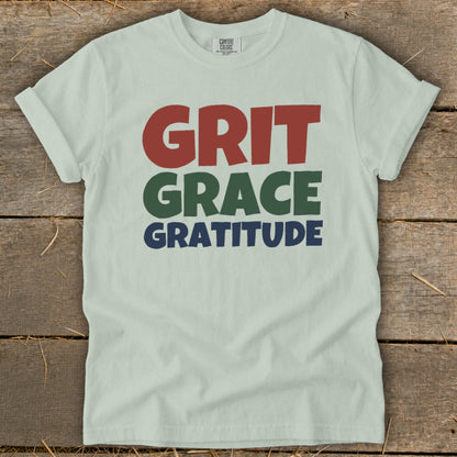Light green t-shirt laid flat on a rustic wooden background, featuring the words Grit, Grace, and Gratitude.