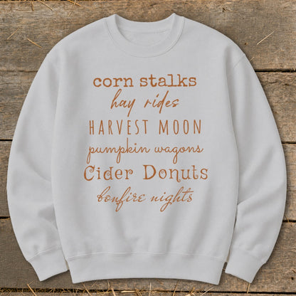 White sweatshirt with fall-themed text: corn stalks, hay rides, harvest moon, pumpkin wagons, cider donuts, bonfire nights.