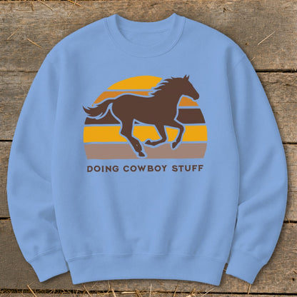 Light blue sweatshirt with a brown horse silhouette running in front of a striped sunset and the text "DOING COWBOY STUFF."