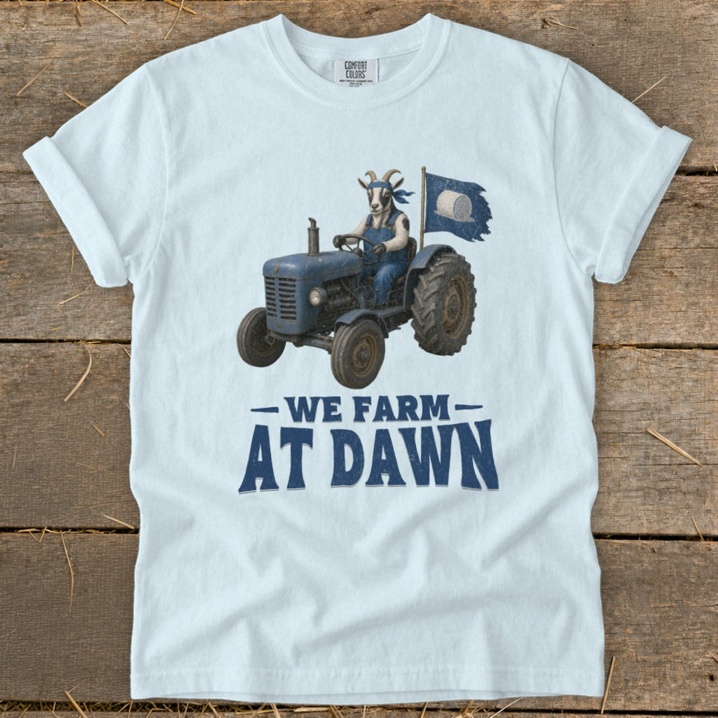 Light blue t-shirt with a goat in overalls driving a blue tractor, "We Farm At Dawn" text.
