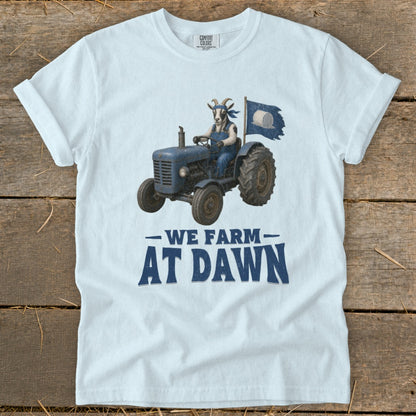 Light blue t-shirt with a goat in overalls driving a blue tractor, "We Farm At Dawn" text.