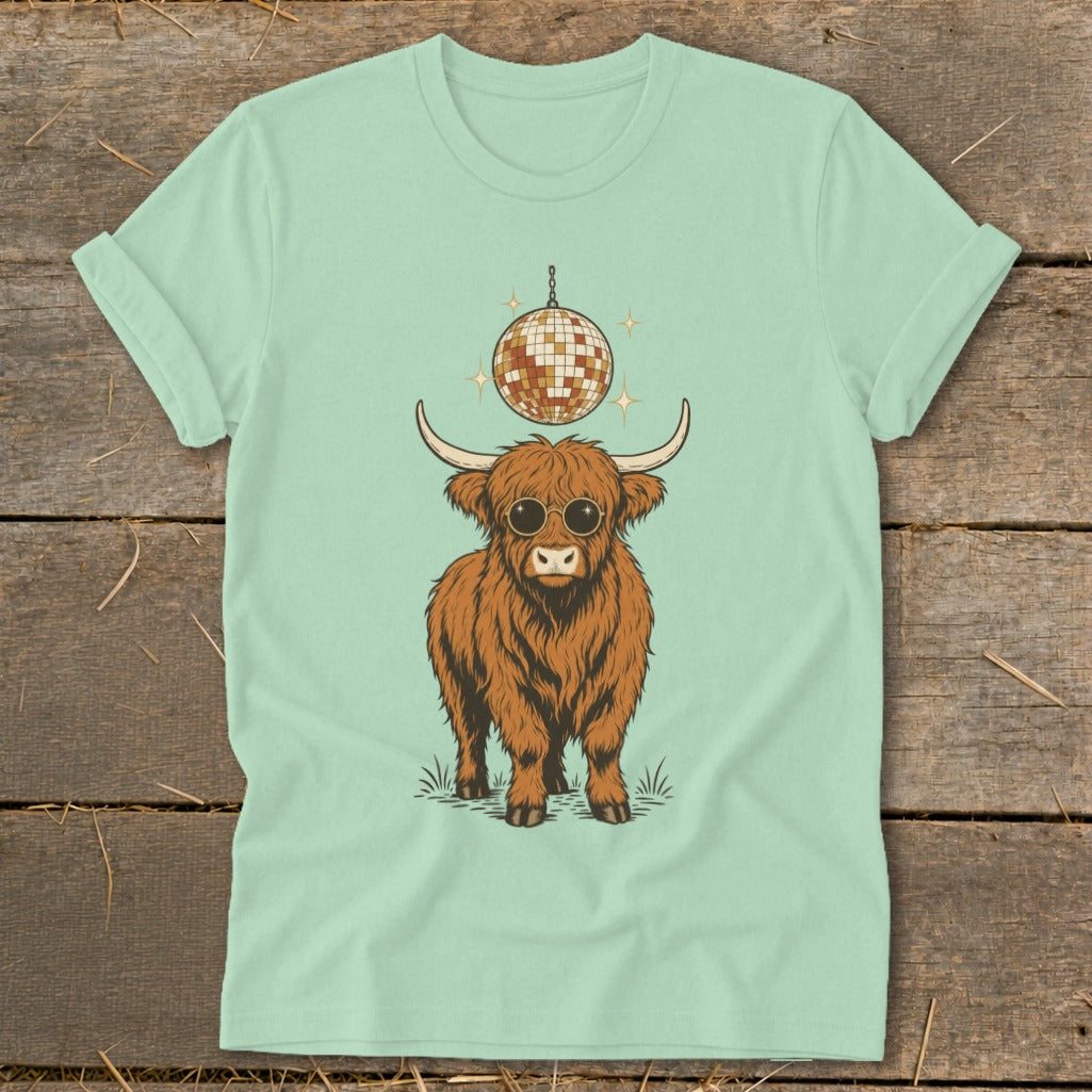 Highland cow wearing sunglasses under disco ball graphic on a light green t-shirt.