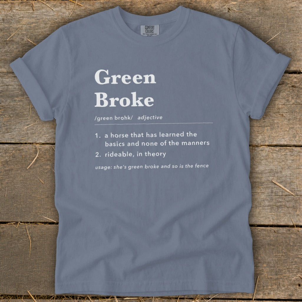 A blue-grey t-shirt lies flat on a wooden surface, featuring the white text definition for Green Broke.