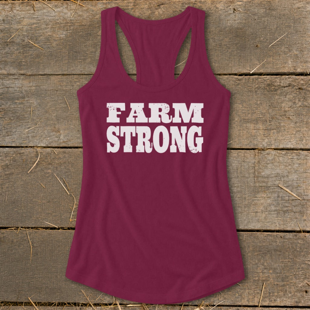 Maroon women's racerback tank top featuring the white distressed text Farm Strong, laid flat on wooden planks.