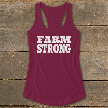 Maroon women's racerback tank top featuring the white distressed text Farm Strong, laid flat on wooden planks.