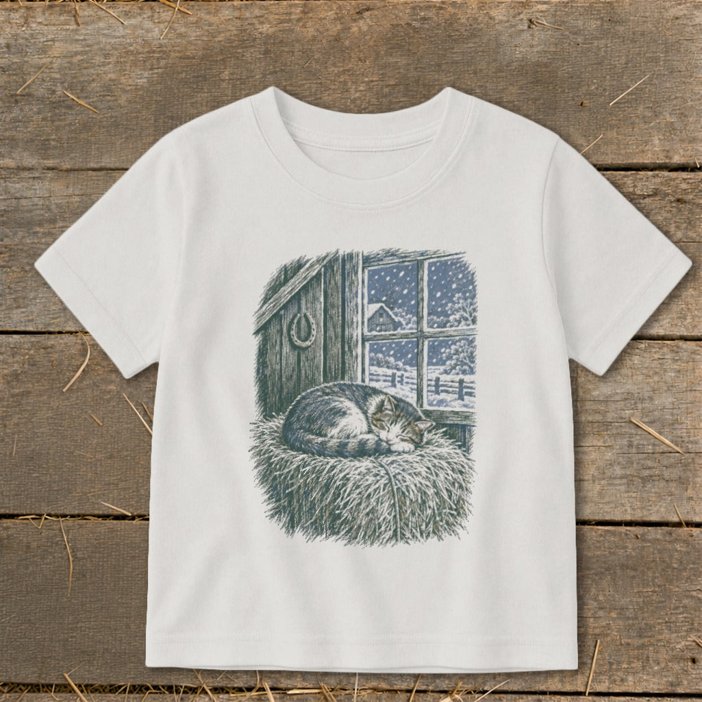 White t-shirt with a drawing of a cat sleeping on hay by a snowy window.