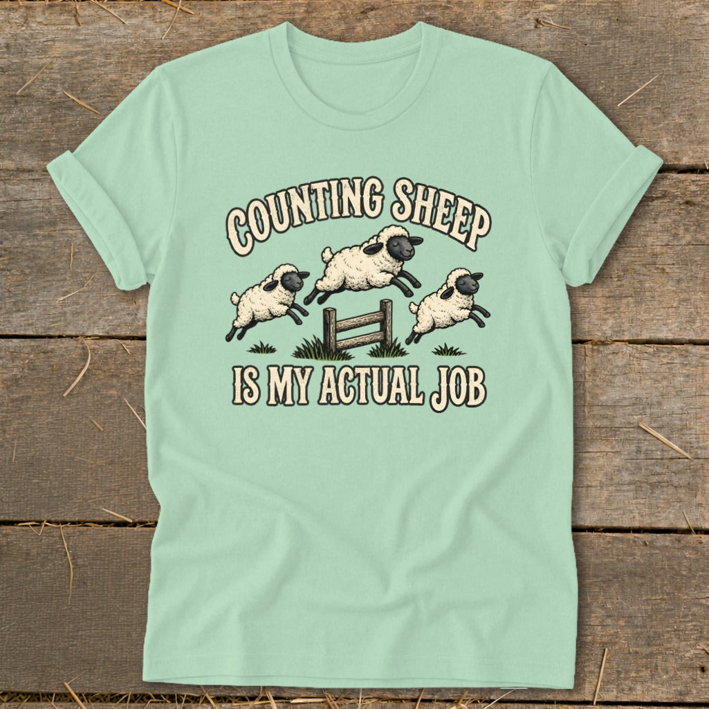 Light blue t-shirt with illustration of three sheep jumping over a fence, text "COUNTING SHEEP IS MY ACTUAL JOB".