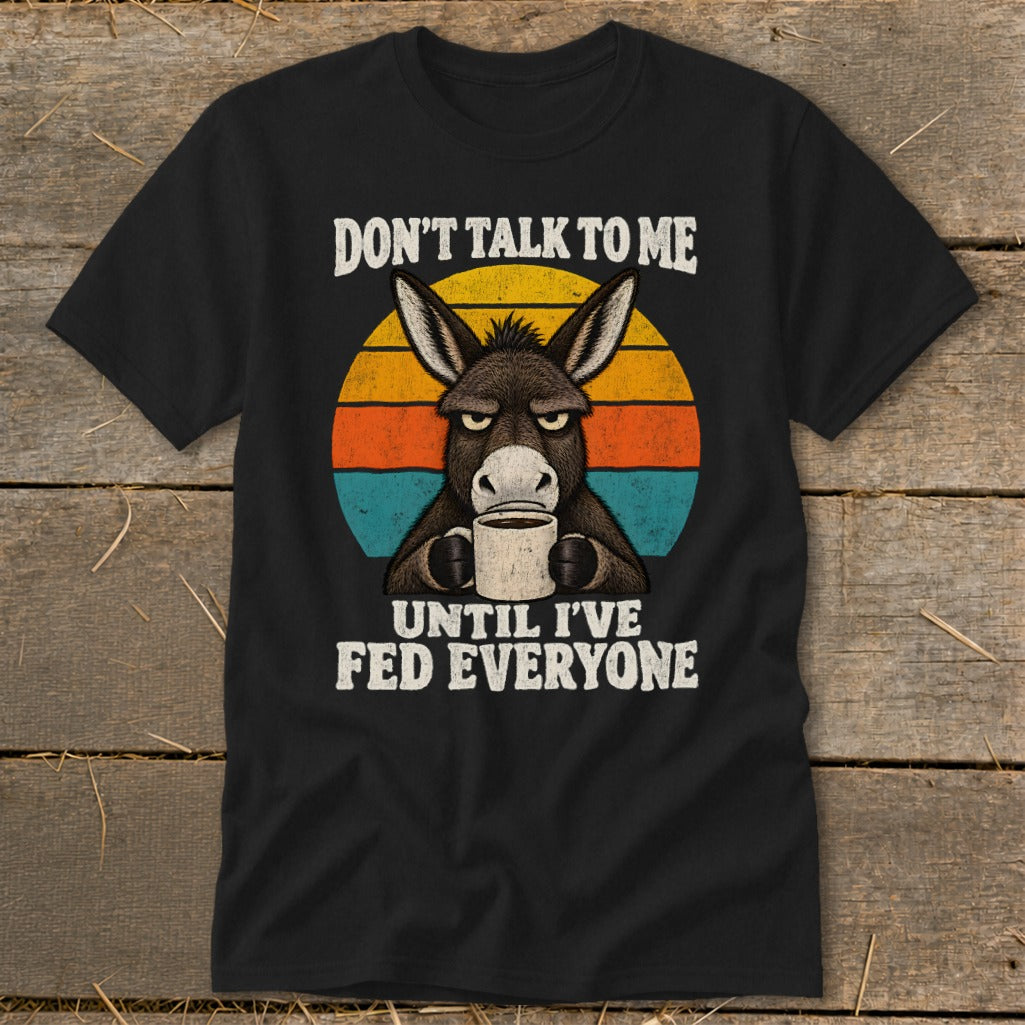 A black t-shirt features a graphic of a grumpy donkey holding a coffee mug, overlaid on a colorful retro sunset circle with the text "DON'T TALK TO ME UNTIL I'VE FED EVERYONE."
