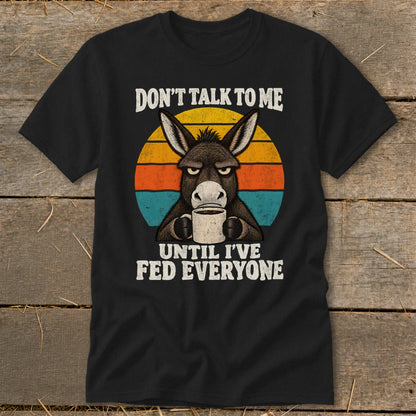 A black t-shirt features a graphic of a grumpy donkey holding a coffee mug, overlaid on a colorful retro sunset circle with the text "DON'T TALK TO ME UNTIL I'VE FED EVERYONE."