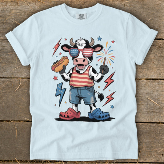 Cartoon cow in patriotic sunglasses holding hot dog and sparkler.